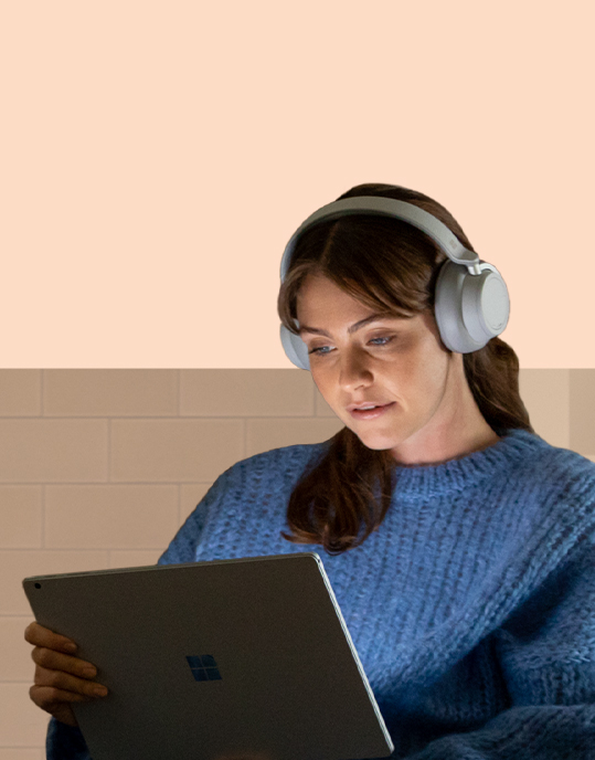 A woman looks at her Surface device that lights up her face while wearing Surface Headphones 2.