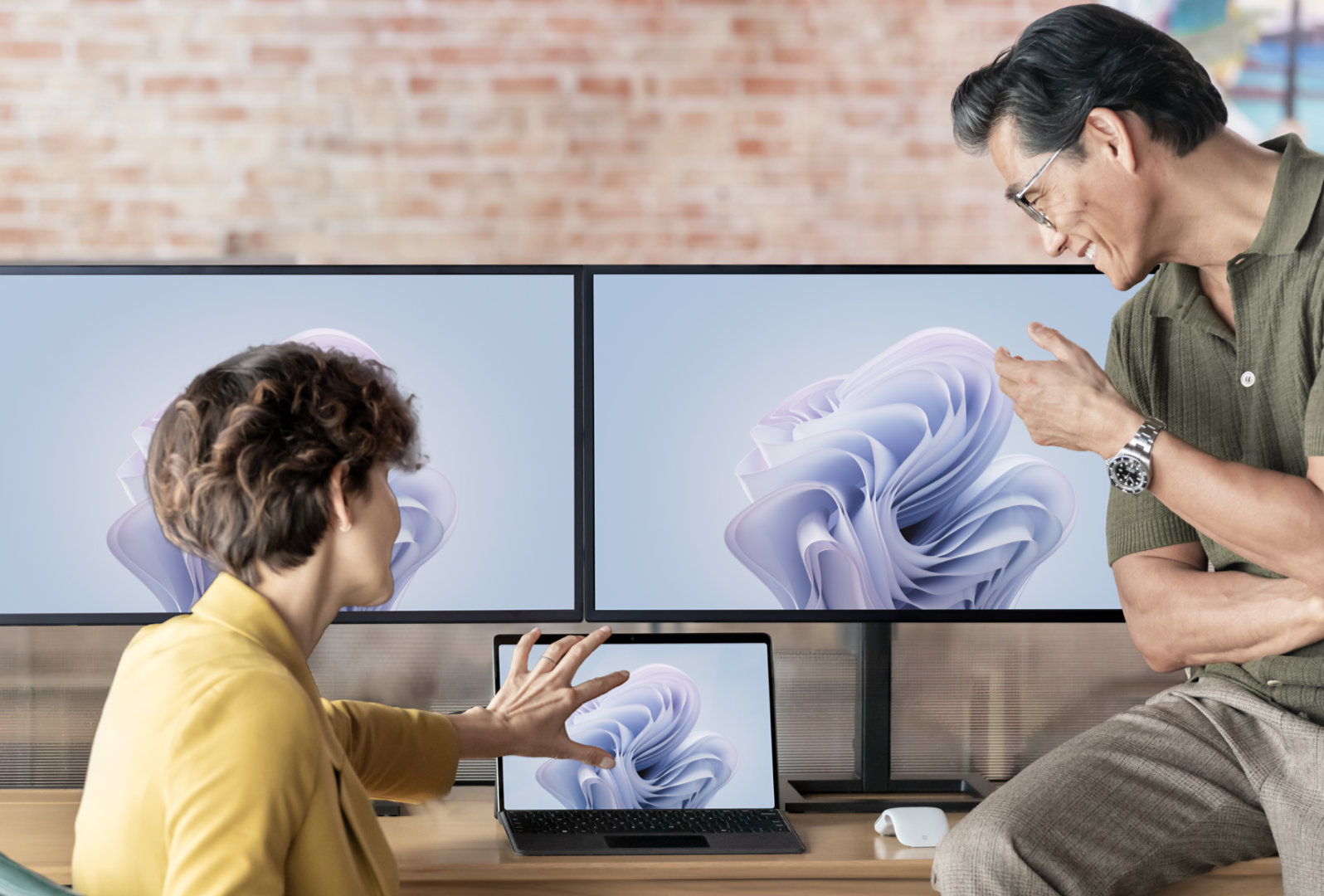 一男一女坐在办公桌前，看着 Surface Pro 10，此设备连接到 2 台显示有 Microsoft 365 应用程序的显示器