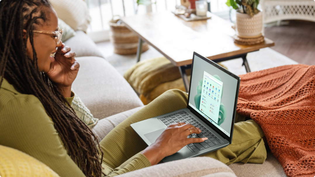 A women sitting on a couch types on a Surface Laptop 5 placed on her lap