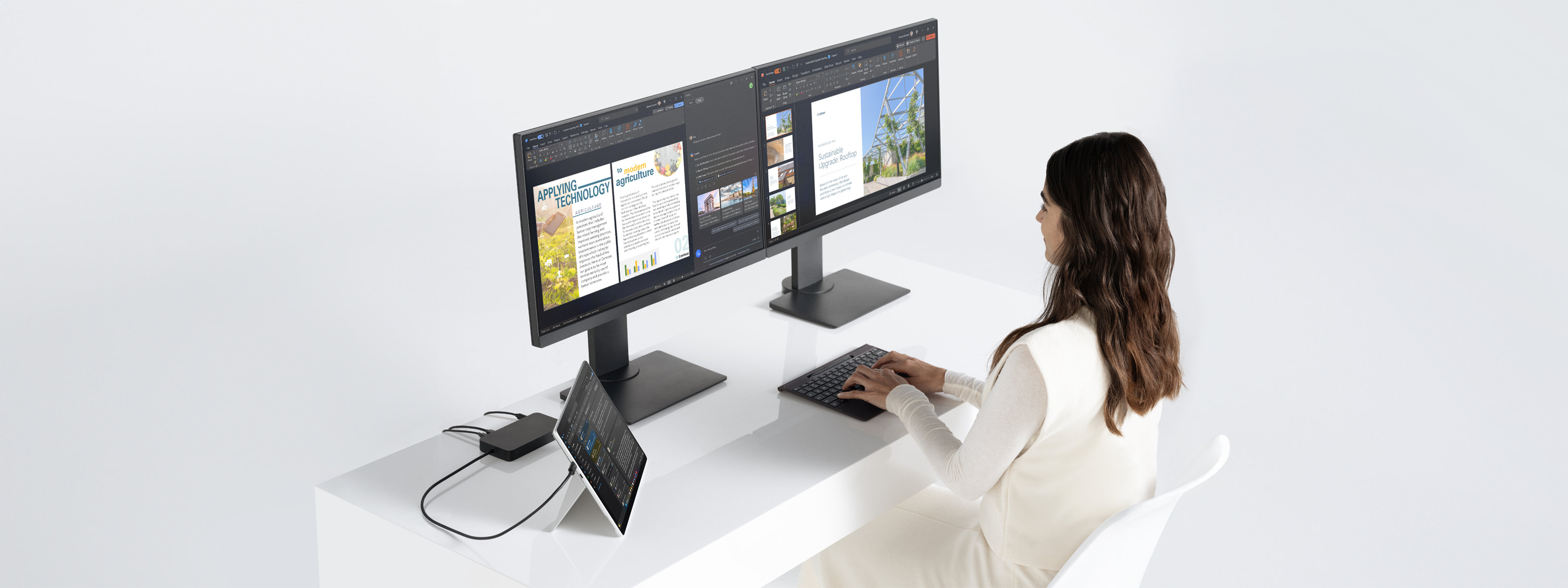 Een vrouw zit aan een bureau met een Surface Pro-apparaat in de kickstandmodus, verbonden met twee monitoren via een dock terwijl ze werkt.