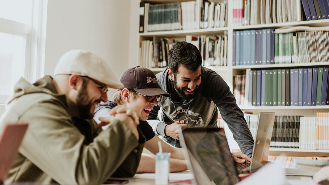 Three men laughing while looking at a laptop in a study room