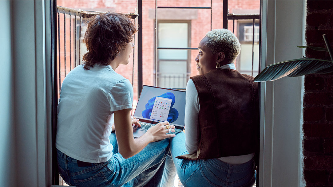 Two Gen Z friends sitting on fire escape balcony with laptop open with Windows 11 start screen and start menu