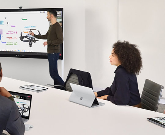 Dwóch współpracowników współpracujących przy użyciu Surface Hub 2S, prowadząc jednocześnie wirtualne spotkanie zespołów na urządzeniu Surface Laptop znajdującym się na sąsiednim biurku