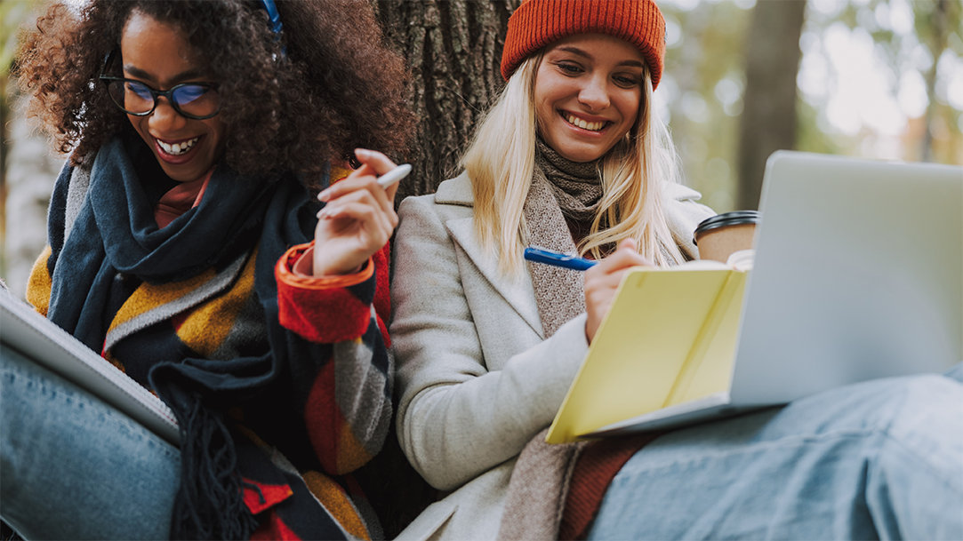 Two college female college buddies studying together outside during a cold afternoon, using Copilot on a laptop