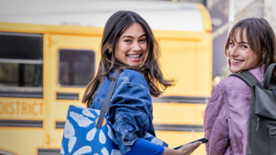 Two female students walking towards a school bus and looking back over their shoulders