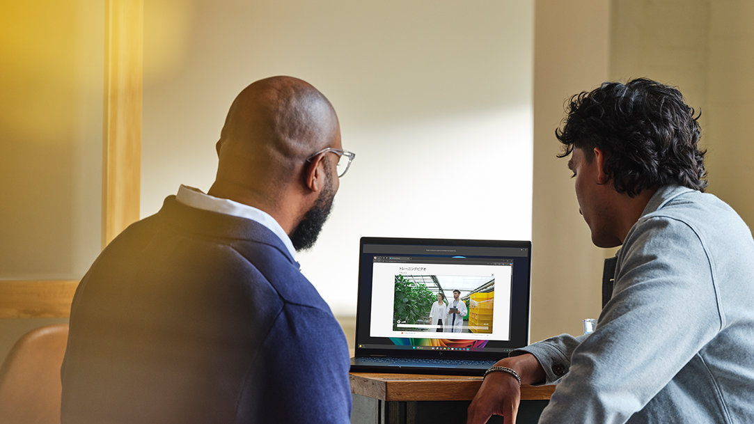 Two people sitting at a desk watching a video on a laptop running Windows 11 Pro, with a window and soft yellow light in the background