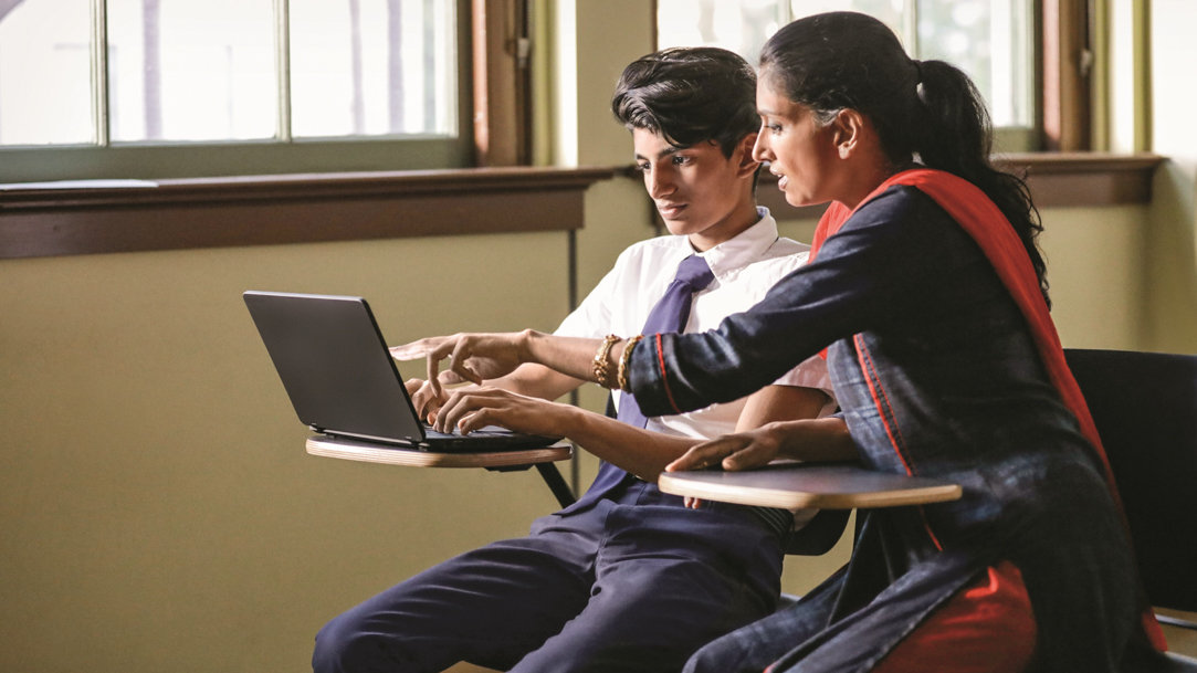 Two people sitting at desks in a classroom and using a laptop together