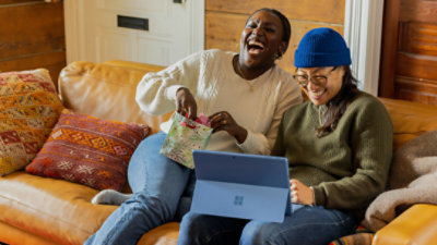 Two women sitting on a couch laughing while looking at a laptop