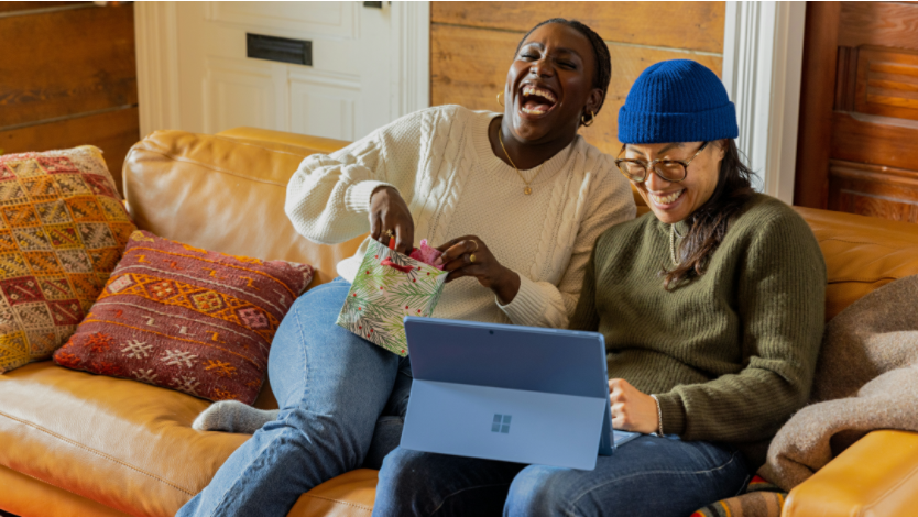 Two women sitting on a couch laughing while looking at a laptop