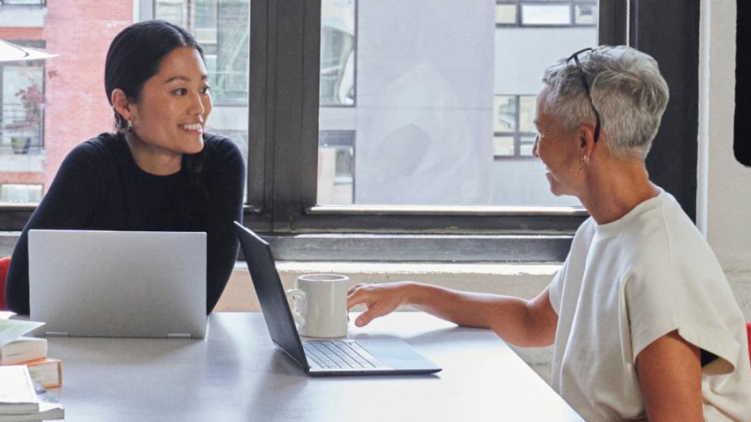 Zwei Frauen, vor denen jeweils ein geöffneter Windows-Laptop steht, unterhalten sich an einem Büroarbeitsplatz.