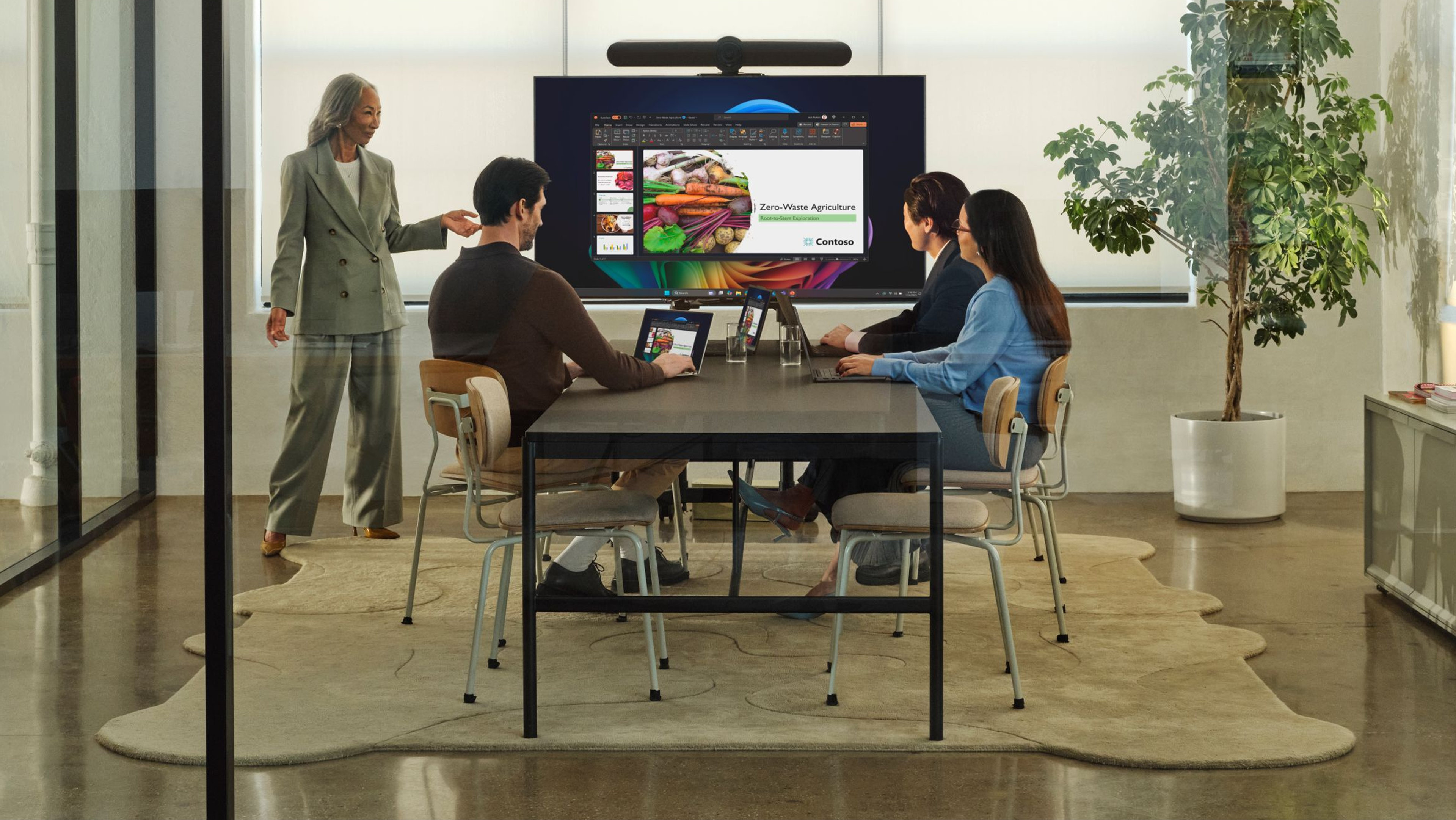 Four office workers with open Windows 11 Pro + Copilot PCs, one standing and three seated at a table, in a modern meeting room with a slideshow projected onto a large screen