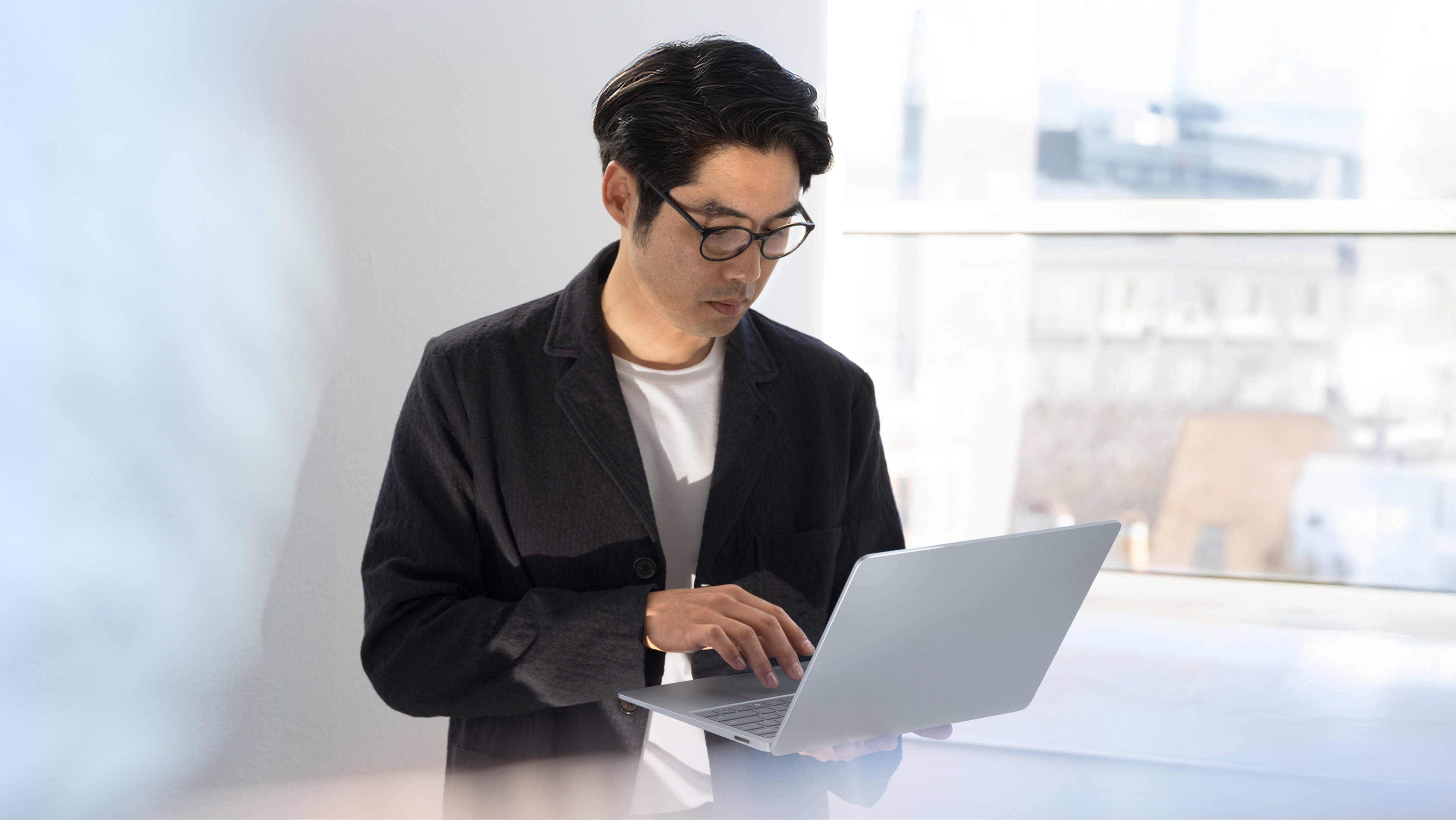 Office worker standing and holding a chrome Surface laptop in an office in front of a window cityscape