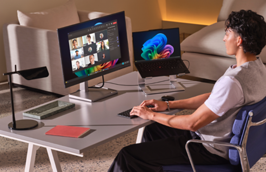 Eine Person sitzt an einem Schreibtisch mit zwei Monitoren, mit einer Videokonferenz auf dem größeren Bildschirm und einer Regenbogenblüte auf dem Laptop, sowie einem beigefarbenen Sofa und Umgebungsbeleuchtung im Hintergrund