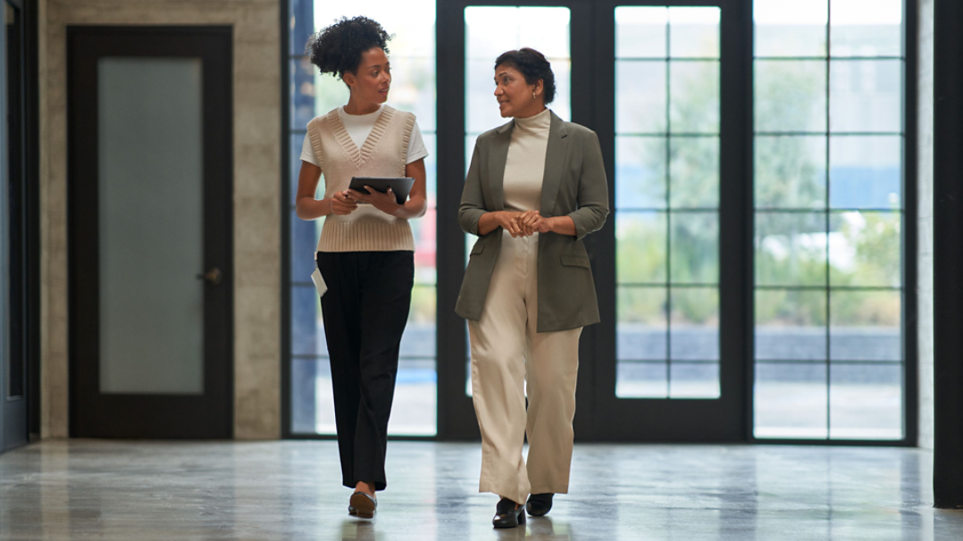 Two people walking together in a brightly lit room with large, paneled windows