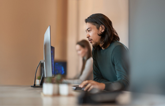 Two people working on monitors at a table, with the focal point on the person in the foreground