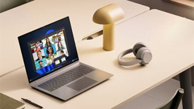 An open laptop sits on a desk alongside a lamp and headphones, with a video call taking place on the screen