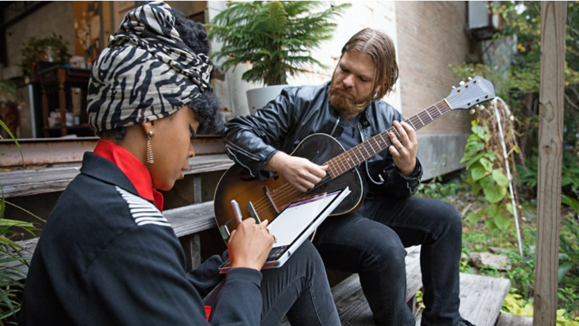 Woman sitting outside on a staircase and taking notes on her Surface device beside a guitarist