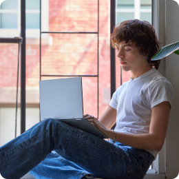 Uma pessoa jovem sentada junto a uma janela aberta enquanto segura num PC