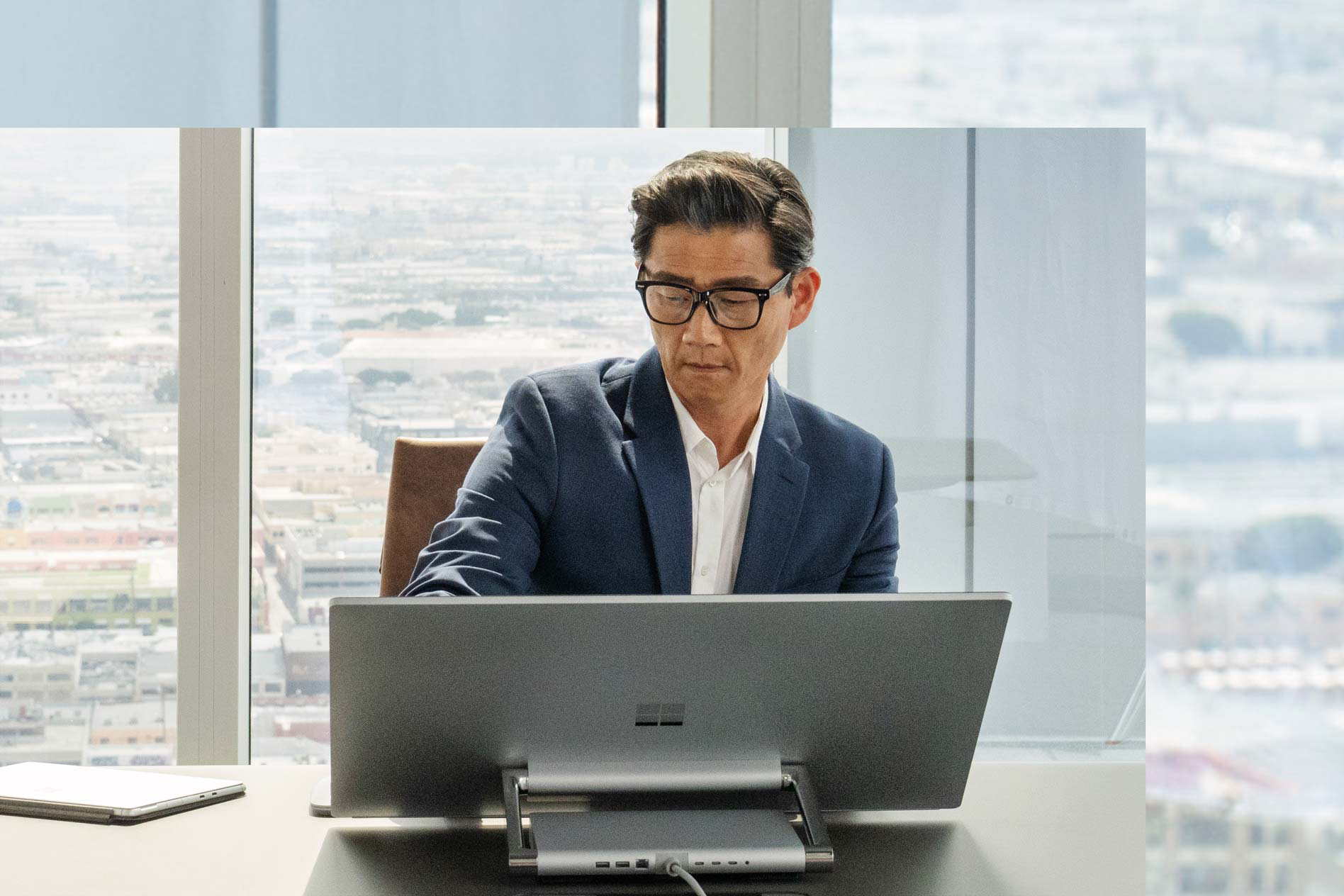 有個男人坐著，窗戶前方的 Surface Studio 2+ 顯示些許城市。