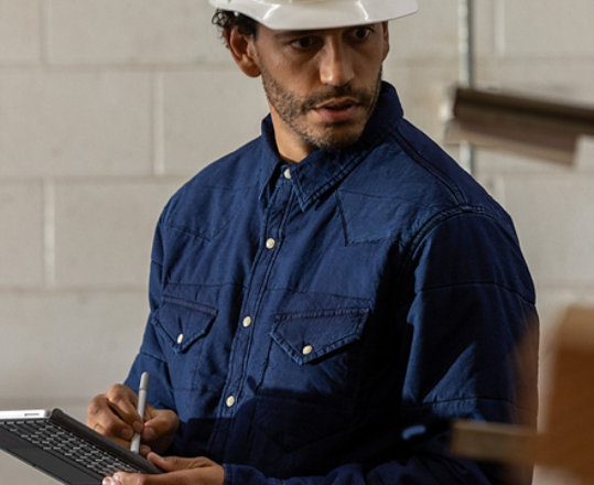 A worker is seen wearing a hard hat in an industrial setting holding a Surface Go 2 in tablet mode