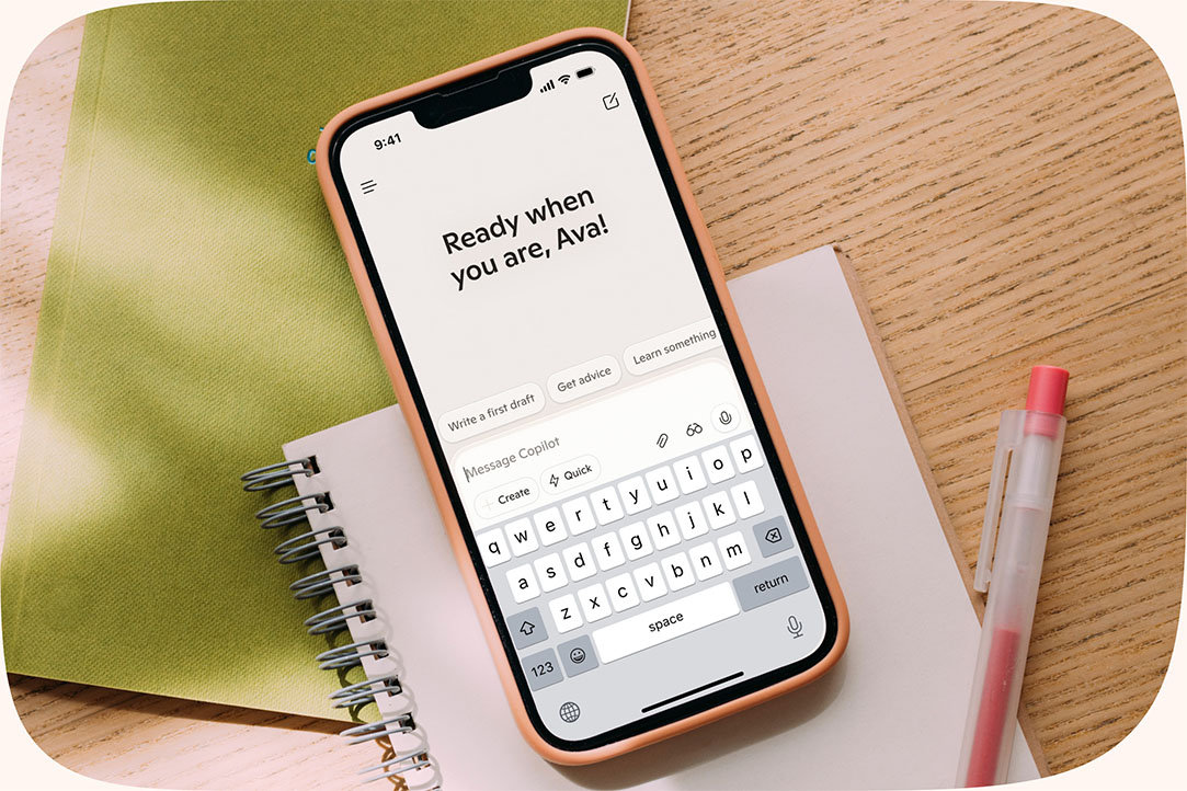 A cellphone on top of a notepad with the screen showing Copilot saying “Ready when you are, Ava!”