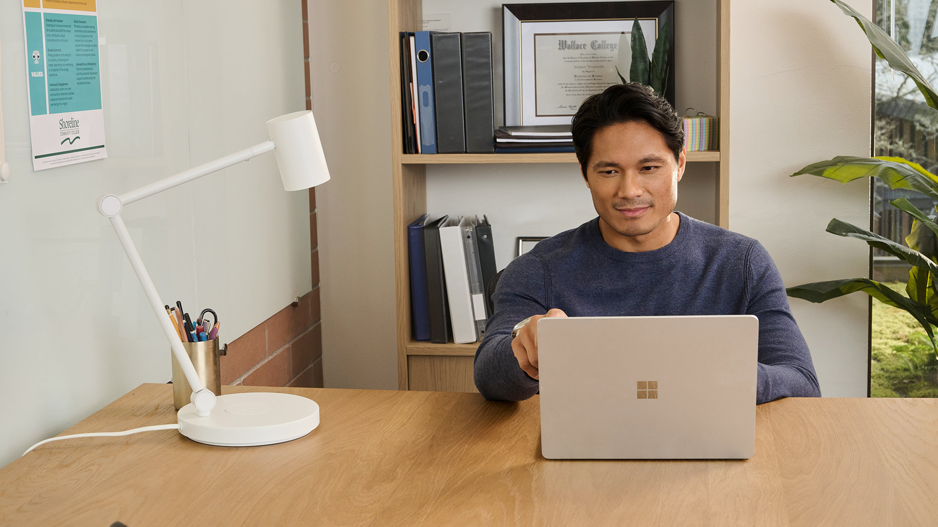 A remote worker sitting at a wooden desk with an open platinum Windows laptop, a desk lamp, and pens, and a bookcase in the background.