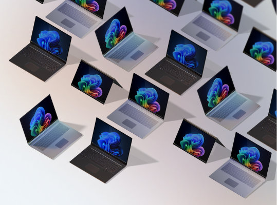 A group of 12 open Windows 11 Pro Edition and Windows PCs for business, some displaying the rainbow bloom image, some displaying the blue rainbow image, all seen from above against a gradient peach background