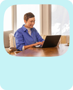 A person sitting at a desk typing on an open Windows laptop with windows in the background
