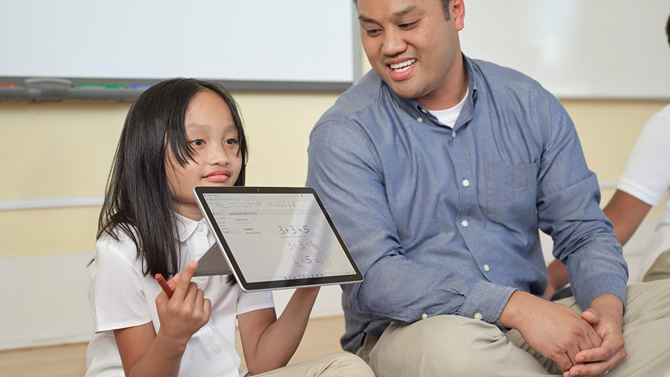 Młoda uczennica pokazująca klasie urządzenie Surface Go i przyglądający się nauczyciel