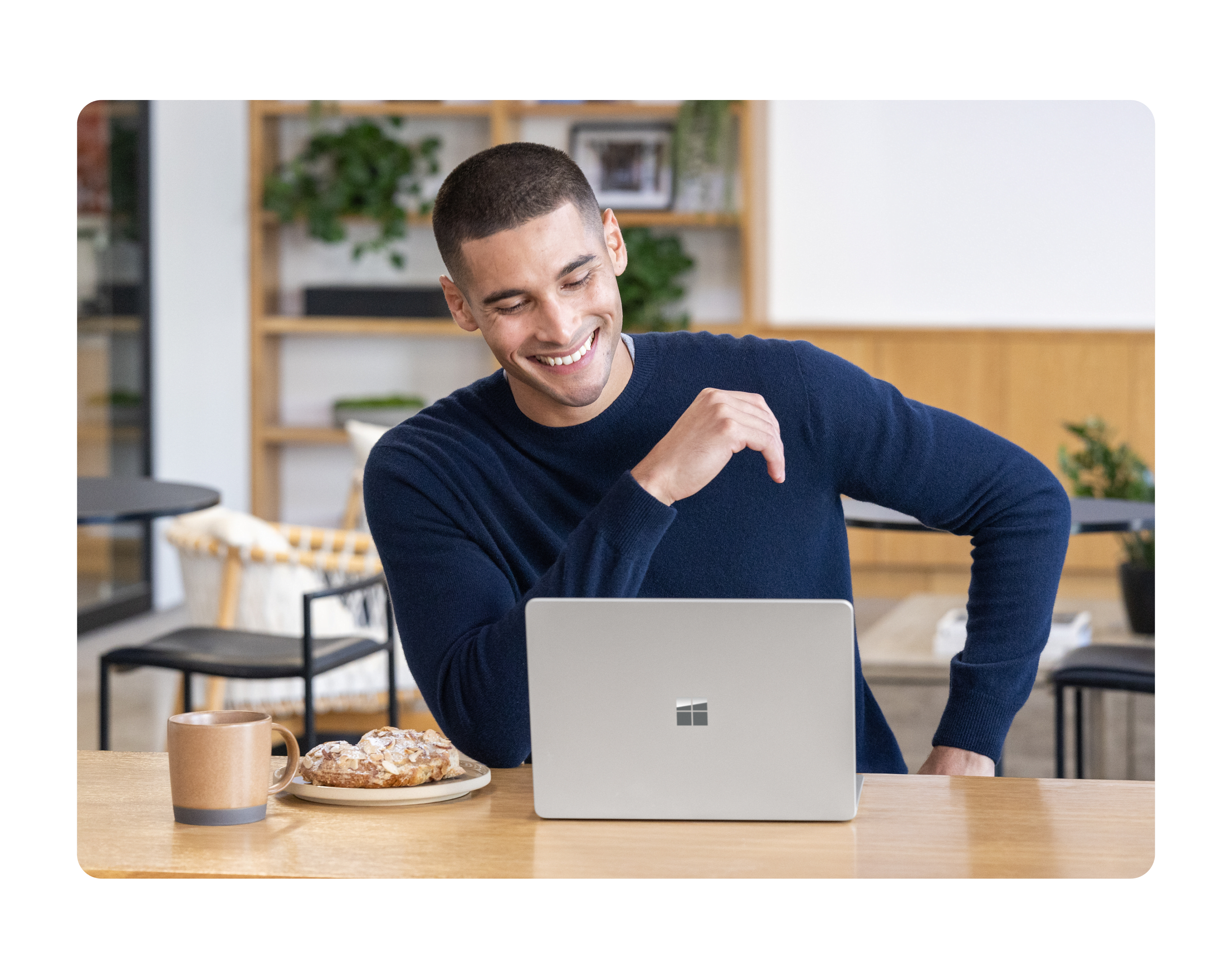 A person smiling working on a Surface laptop alongside a coffee mug and pastry on a wood table