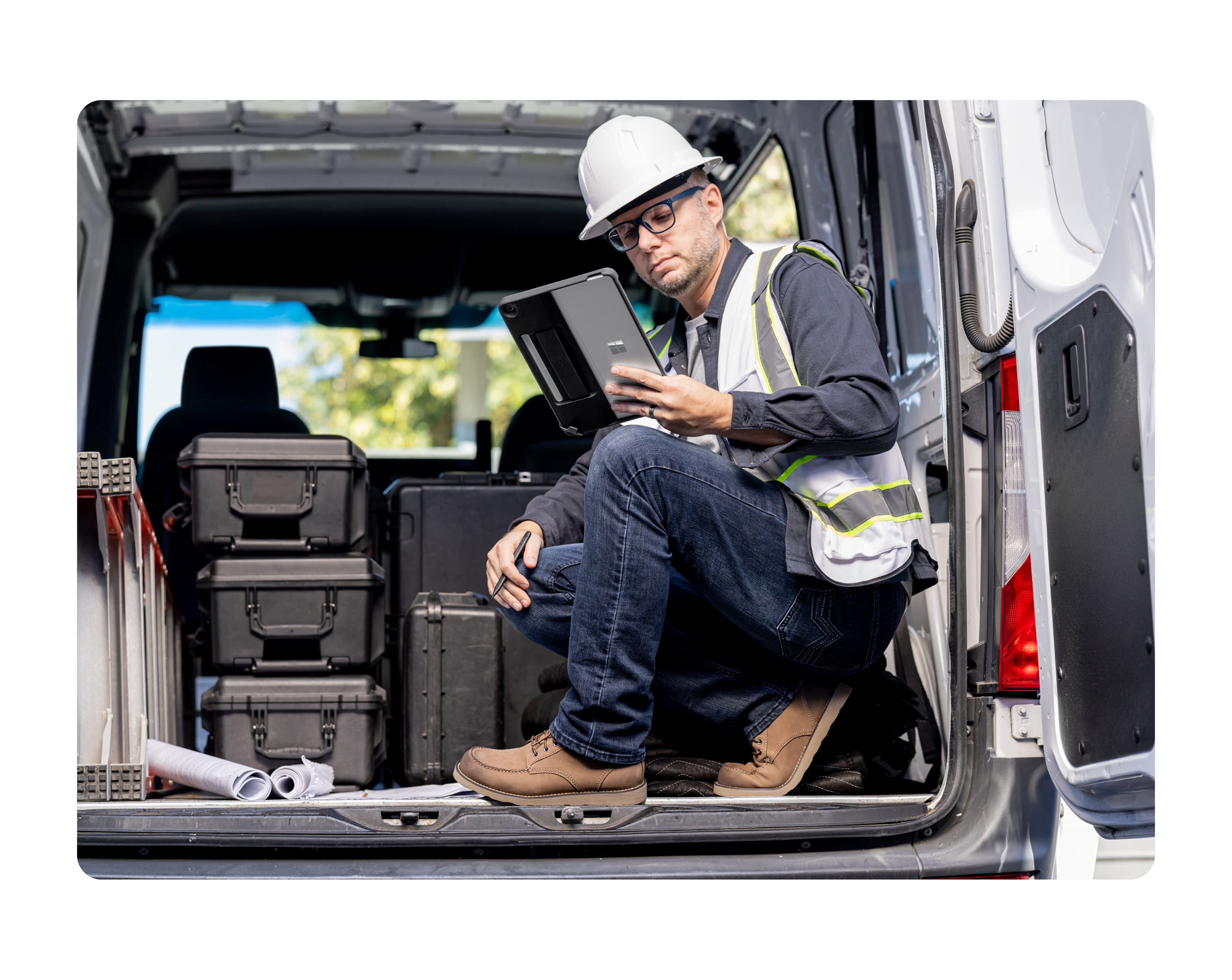 A person in a construction hat and vest sits in the back of a van working on a Surface 2-in-1 device