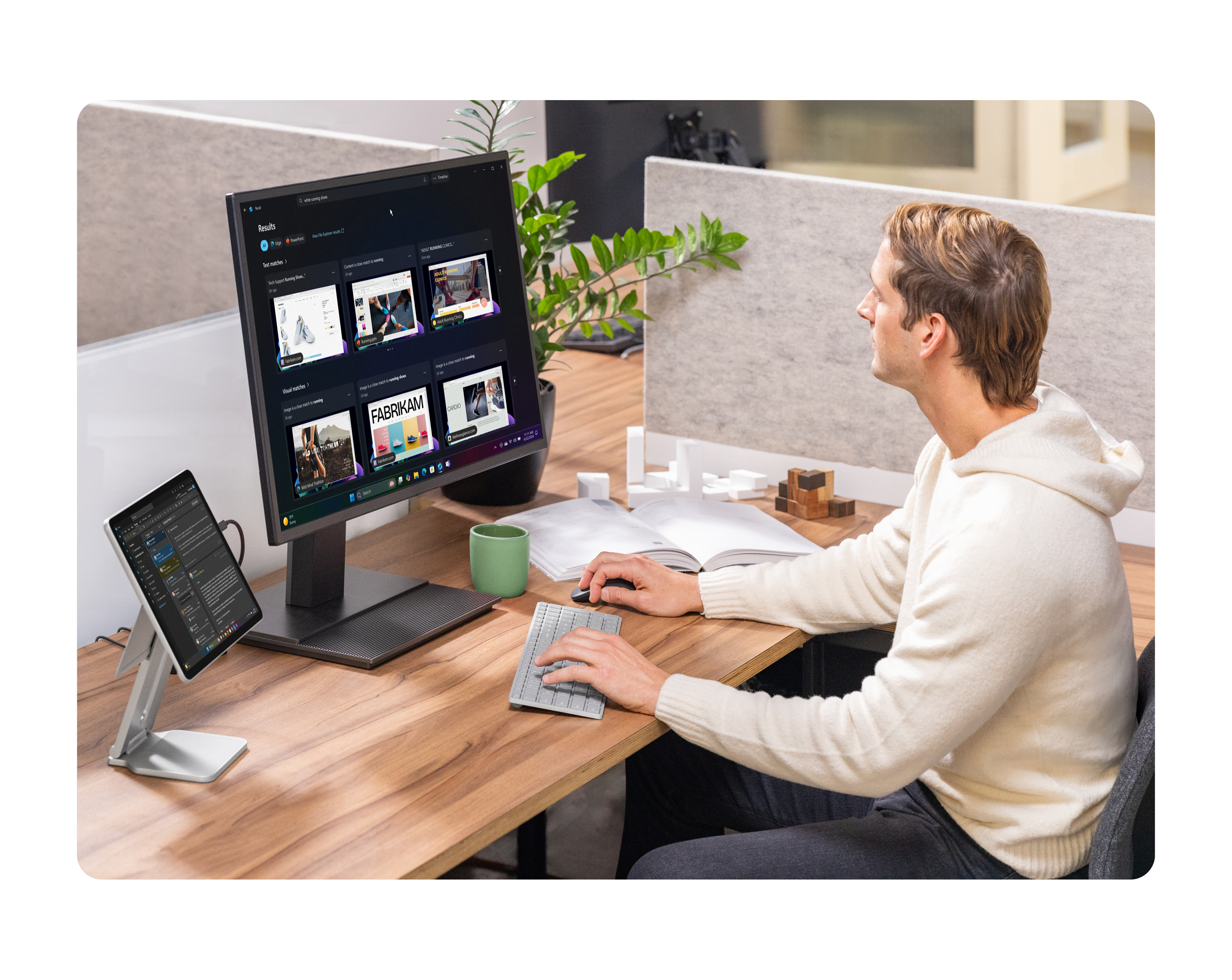 A person working on a computer at a wood desk in a cubicle alongside a 2-in-1 device, open notebook, and plant.