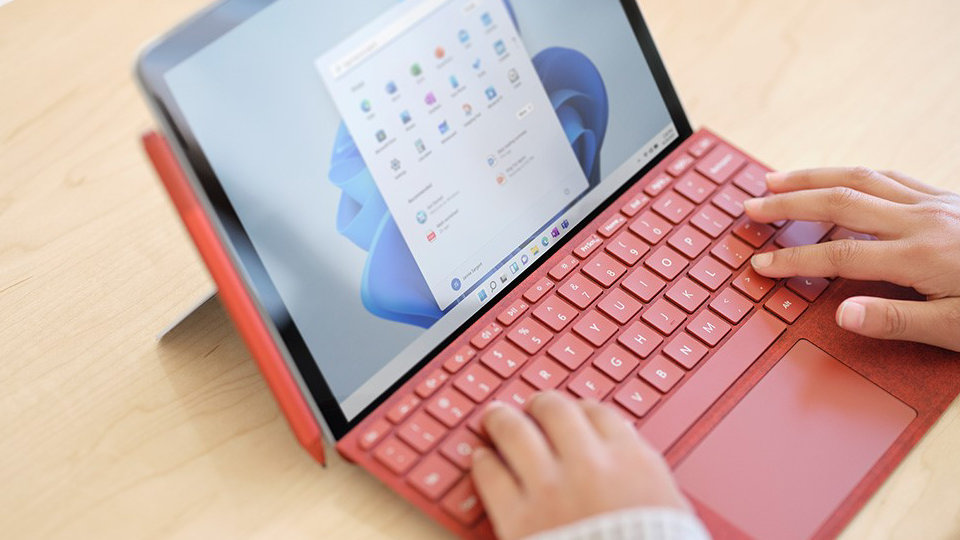 Dwie ręce piszące na klawiaturze Surface Type Cover w kolorze czerwonego maku