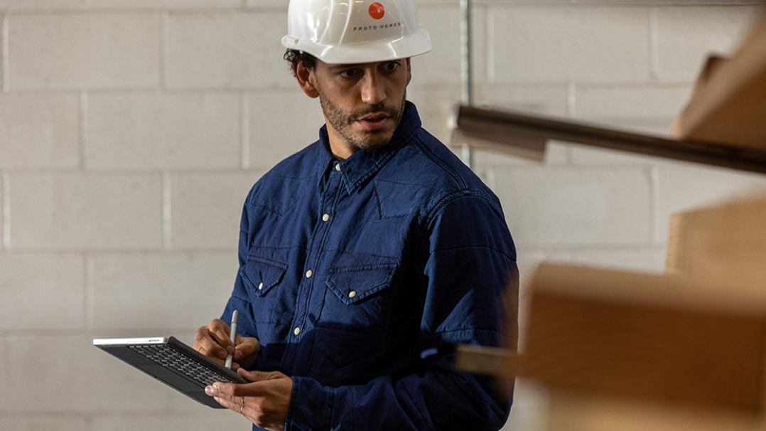 A worker is seen wearing a hard hat in an industrial setting holding a Surface Go 2 in tablet mode