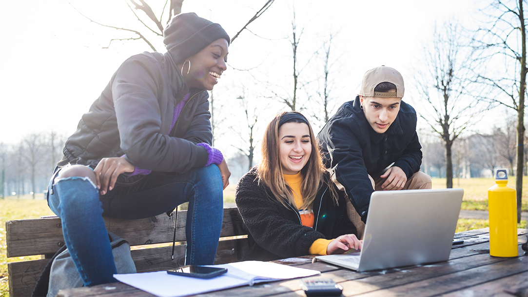 Three high school students sitting at a bench and table in a park, gathered around and using a laptop