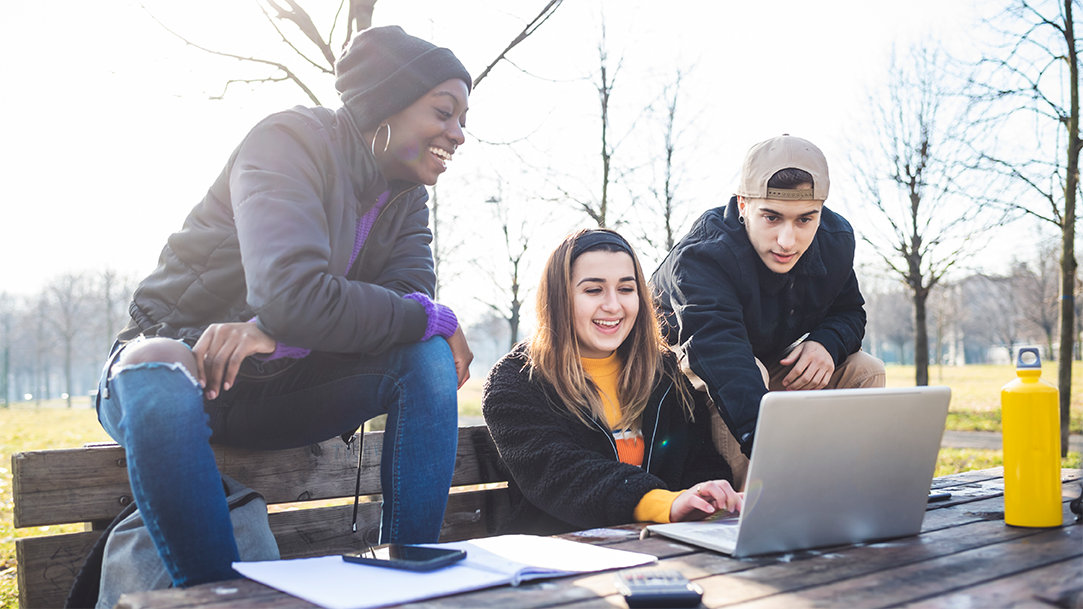 Three high school students sitting at a bench and table in a park, gathered around and using a laptop