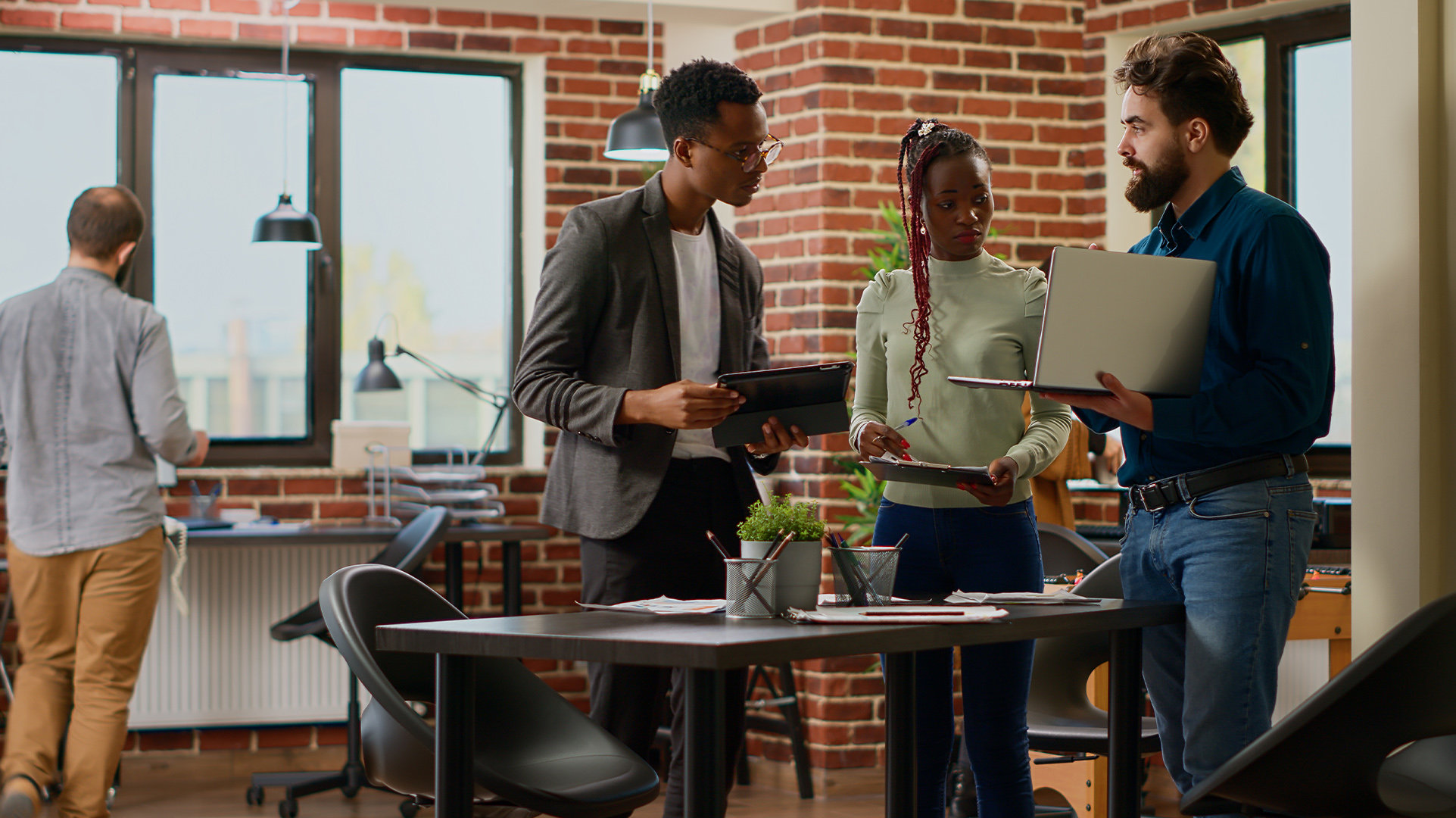 A modern, open office setting with four standing office workers, three of them meeting together over a desk, each holding a Windows 11 Pro device discussing onscreen information