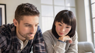 家のリビングで、1人の男性と1人の女性がソファーに並んで座り、会話しています。
