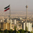 Cityscape of Tehran with Milad Tower in the background and a large Iranian flag in the foreground.