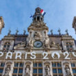A historic building featuring ornate architecture with statues and a central clock. Two flags—France and the European Union—fly atop the central tower. Large white letters spelling 'PARIS 2024' are displayed across the front, referencing the Olympic Games.