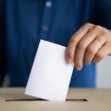 Person in a blue shirt placing a white paper into a ballot box.