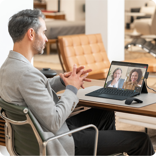 A man is sitting on a chair with a laptop on desk and in a video call with two people.