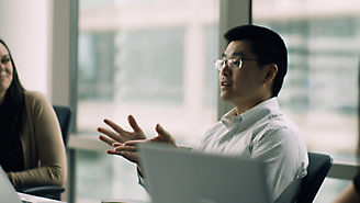 En man med glasögon gestikulerar när han pratar invid en bärbar dator i kontorsmiljö