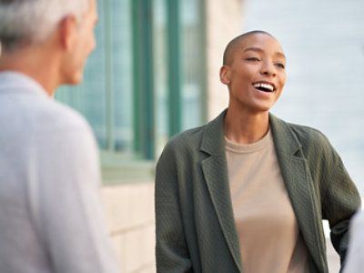 Duas pessoas conversando do lado de fora, uma delas sorrindo. A pessoa em foco está vestindo uma jaqueta verde e uma camisa bege.
