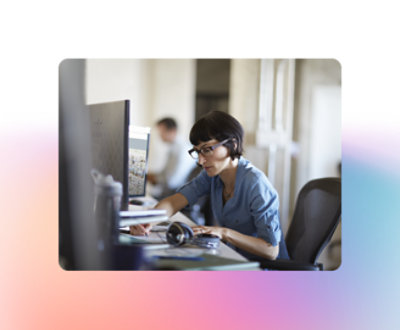Una persona con gafas y camisa azul está sentada en un escritorio, trabajando en un ordenador, con diversos materiales de oficina a su alrededor