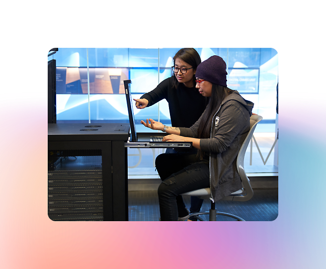 Two people working together at a computer station; one is seated and typing, while the other stands