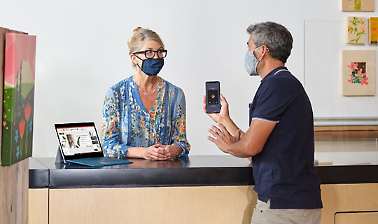 A customer and a receptionist wearing masks and having a conversation at a front desk of a business. There is a Surface device displaying a PowerPoint presentation and the customer is holding up their mobile phone displaying a Teams call in progress