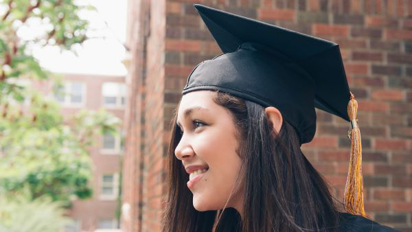 Female student standing outside wearing a graduation ceremony cap and gown.