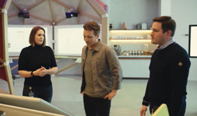 Still image from video showing three people standing together looking down at a large screen.