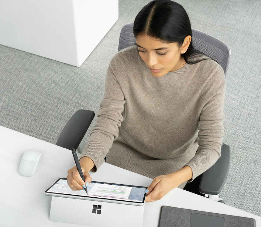 A person uses a Surface Slim Pen for Business to write on the touchscreen of a Surface device, surrounded by other accessories, such as a Surface Arc Mouse and a Surface Pro Keyboard.